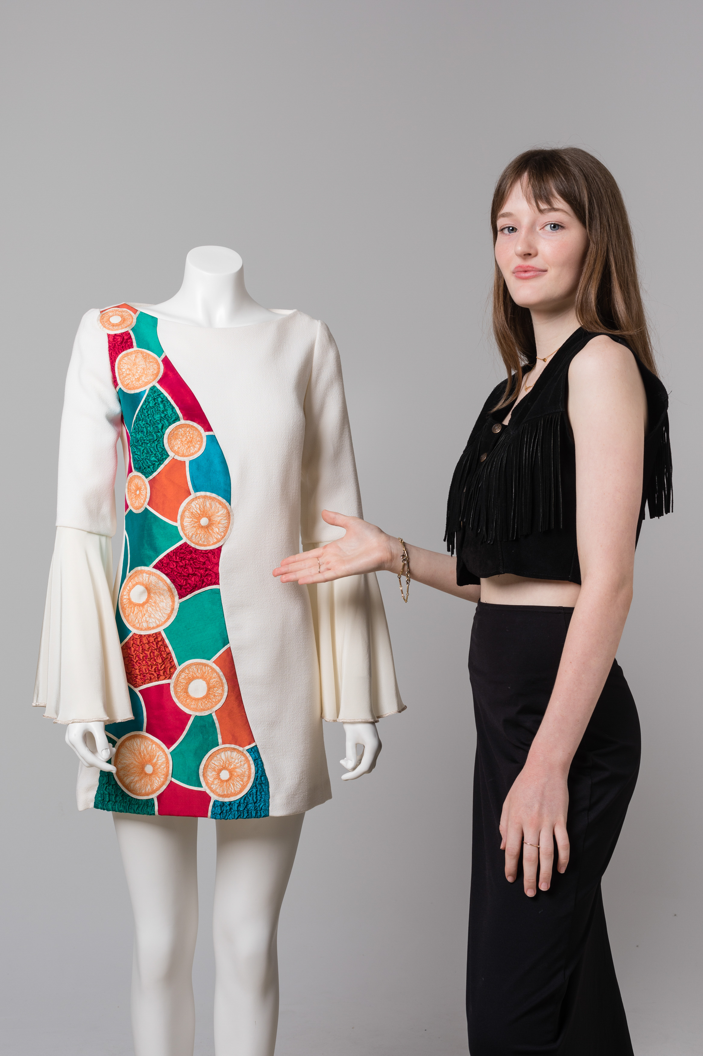 A young woman gesturing to a mannequin beside her. The mannequin wears a long sleeved dress with a colourful pattern down one side and white fabric on the other.